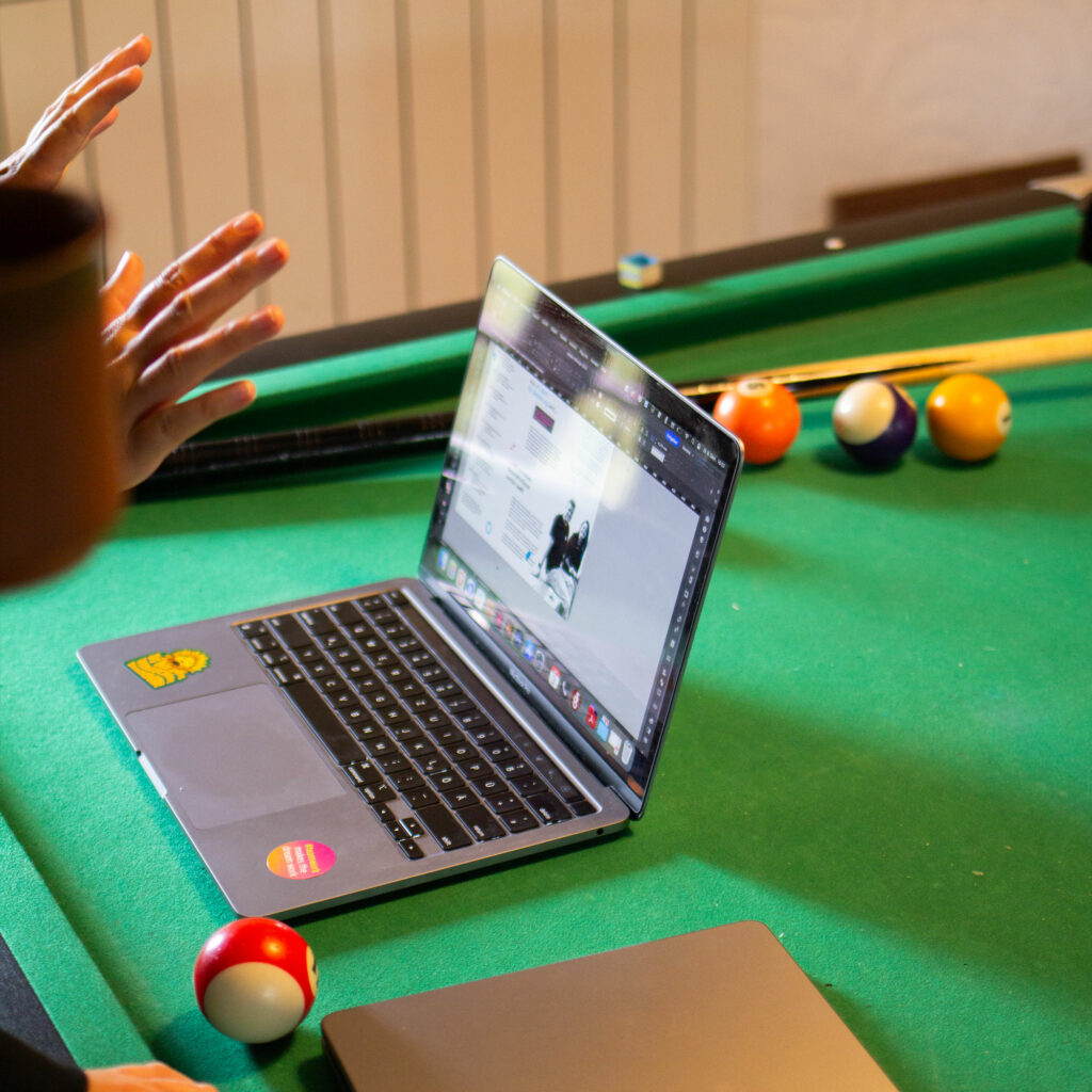Ein Laptop steht auf einem Billardtisch, auf dem Tisch zu sehen sind Billardkugeln und ein Billardqueue.