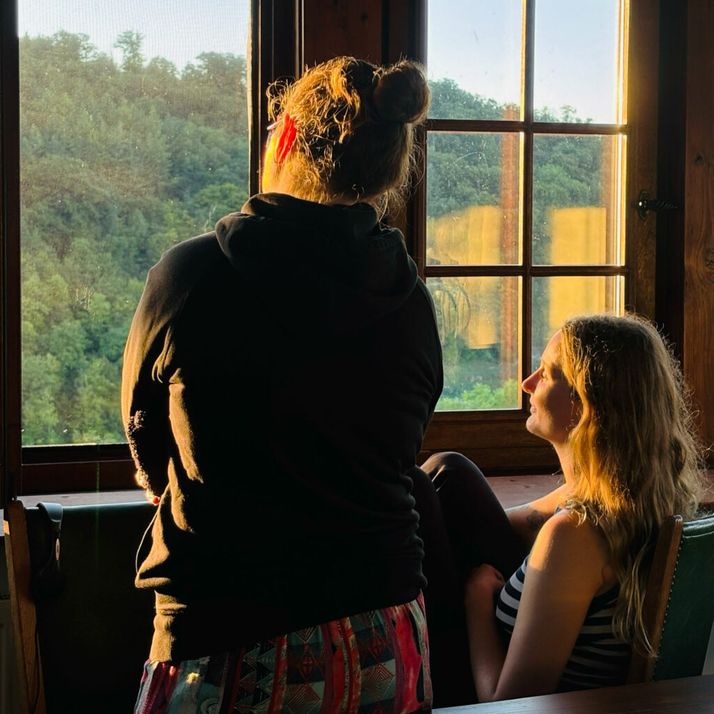 Zwei Mitarbeiterinnen von jo's büro blicken aus einem alten Holzfenster und genießen die Sonnenstrahlen der Abendsonne.