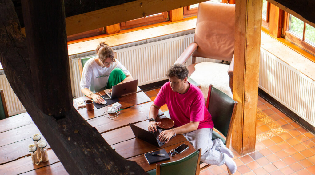 Eine Mitarbeiterin von jo's büro und Jo selbst sitzen vor ihren Laptops an einem Holztisch und arbeiten.